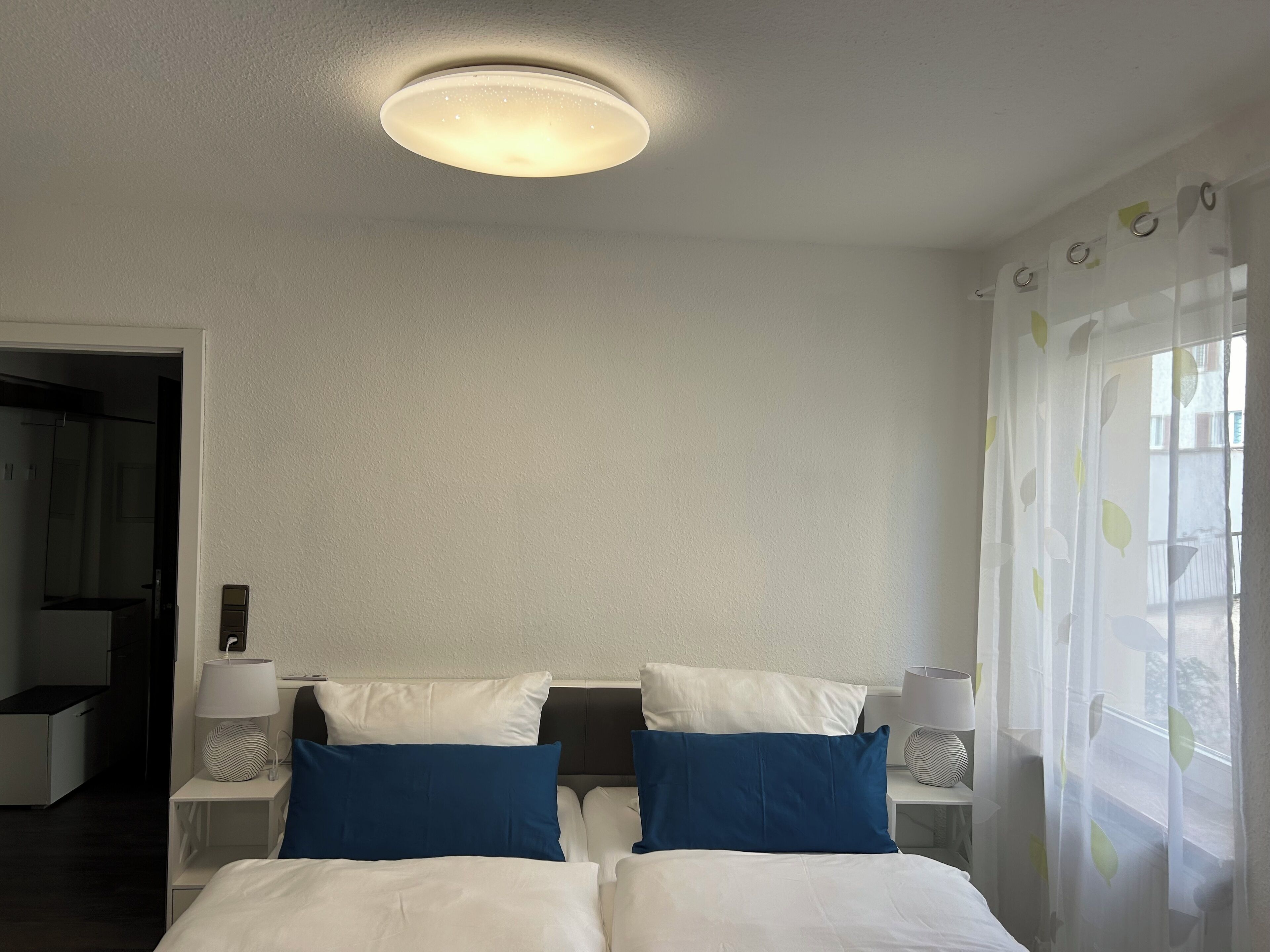A hotel room with a bed, two bedside tables with lamps, a ceiling light, and a window with sheer curtains.