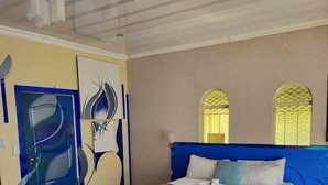 A hotel room with a bed, a red chair, a blue chair, and a door with abstract wall art. - AT BLACK WOOD GUEST HOUSE  (Pretoria)