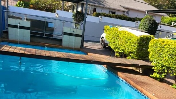 A swimming pool with a wooden deck, surrounded by greenery and a house in the background. - AT BLACK WOOD GUEST HOUSE  (Pretoria)