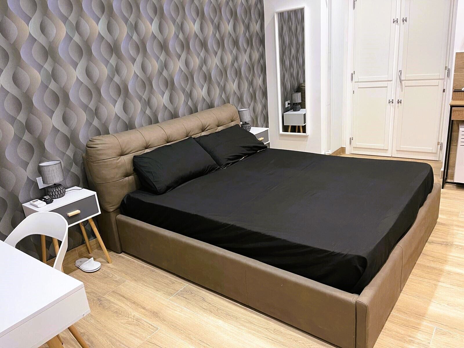 Une chambre avec une tête de lit capitonnée, un lit avec une literie noire, une table de chevet avec une lampe et un petit bureau avec une chaise.