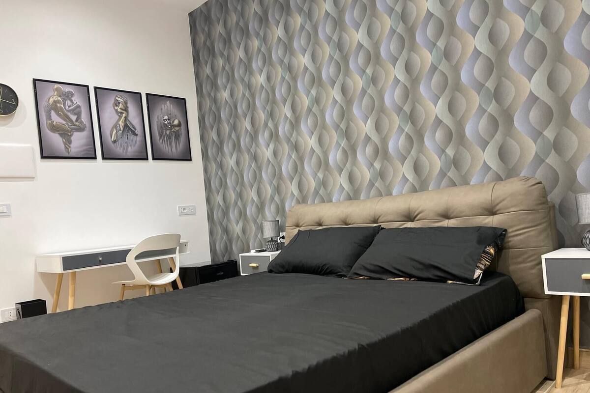 Une chambre moderne avec une tête de lit capitonnée, un lit avec une literie noire, un bureau avec une chaise et un mur à motifs.
