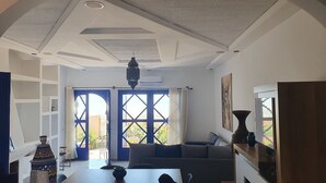 Ein Wohnzimmer mit einer Couch, einem Couchtisch und einer dekorativen Vase. Es gibt ein großes Fenster mit blauen Rollläden und einen Blick ins Freie.