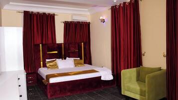 A hotel room with a large bed, red curtains, a green armchair, and a white dresser.