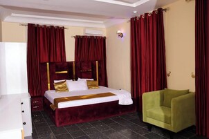 A hotel room with a large bed, red curtains, a green armchair, and a white dresser.