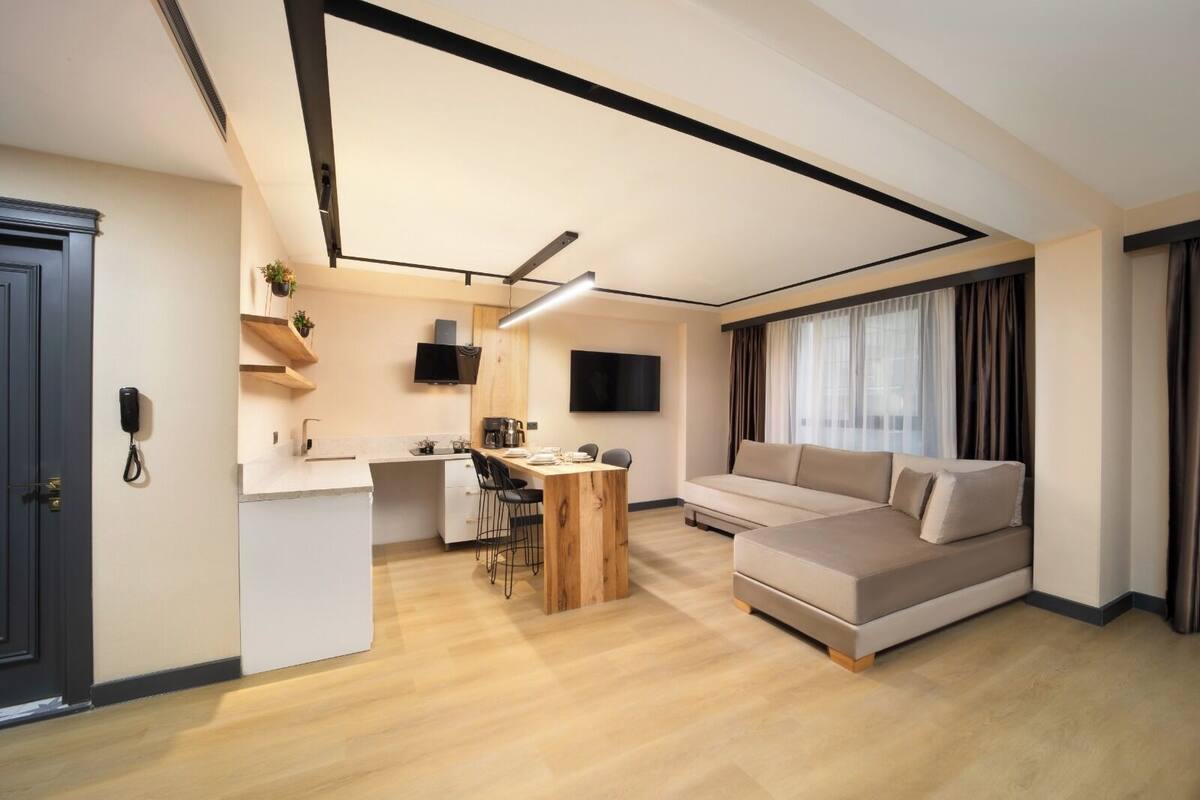 A modern living room with a sofa, a dining area with a table and chairs, a kitchen area, and a TV mounted on the wall.