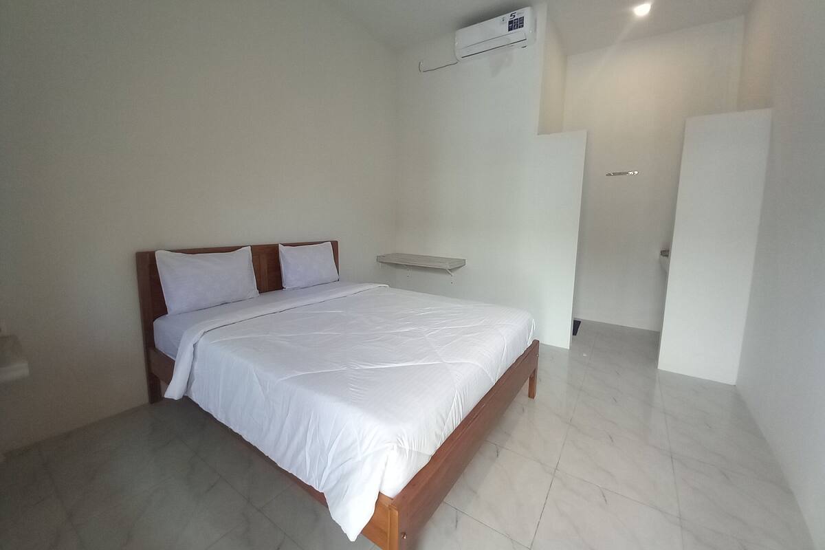 A bedroom with a bed, a white wall, a door, and an air conditioning unit.