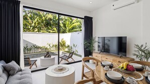 Una sala de estar moderna con un televisor grande, comedor y vista a un jardín exuberante a través de ventanales.