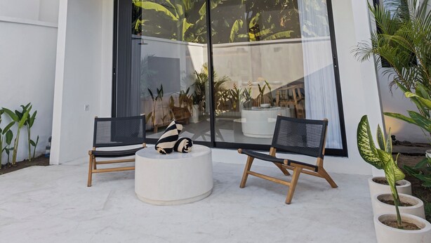 Una casa moderna de dos pisos con techo de madera, ventanas de vidrio amplias y un patio con sillas y plantas en macetas.
