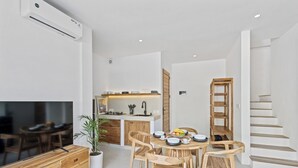 Una cocina moderna con armarios blancos, una mesa de comedor de madera y sillas. Hay un televisor a la izquierda, una planta a la derecha y una escalera que sube a otro nivel.