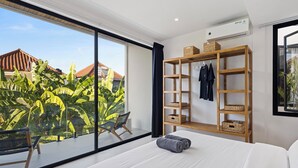 Un dormitorio moderno con un ventanal grande, un armario de madera, una cama con ropa de cama blanca y vistas a un jardín tropical.
