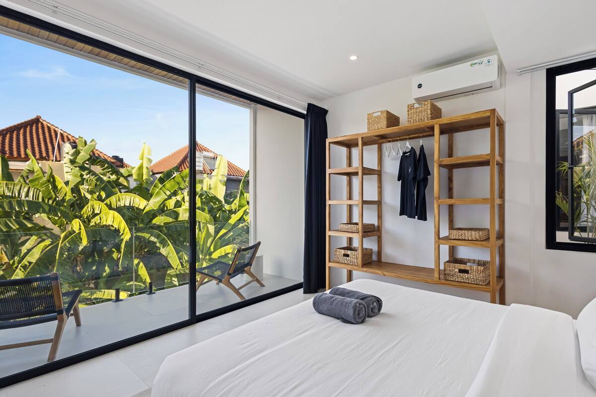 A modern bedroom with a large window, a wooden wardrobe, a bed with white linens, and a view of a tropical garden.