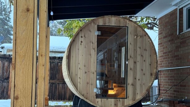 A wooden sauna structure with a glass door, situated outdoors with a brick building in the background.