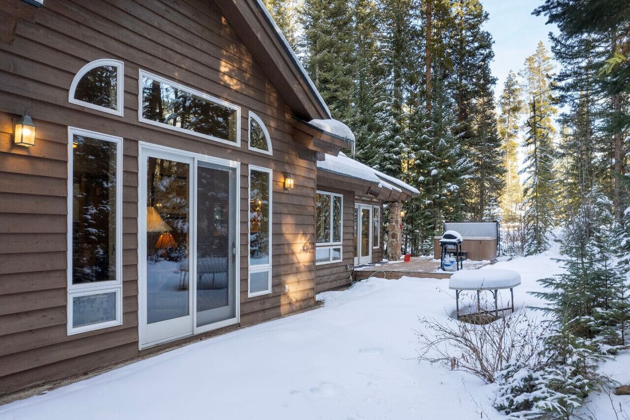 Une cabane en bois dotée de grandes fenêtres, d’un espace extérieur aménagé et couvert, et surplombant un paysage enneigé.