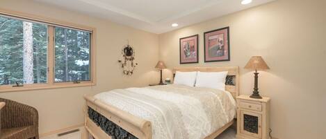 A bedroom with a large window, a bed with a wooden headboard, two bedside tables with lamps, and a wall-mounted dreamcatcher.