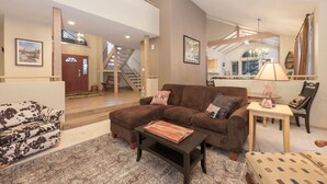 Amplia sala de estar con un sofá seccional marrón, un sillón estampado y una mesa de centro de madera. Una escalera conduce a un nivel superior, y se aprecia un comedor al fondo.