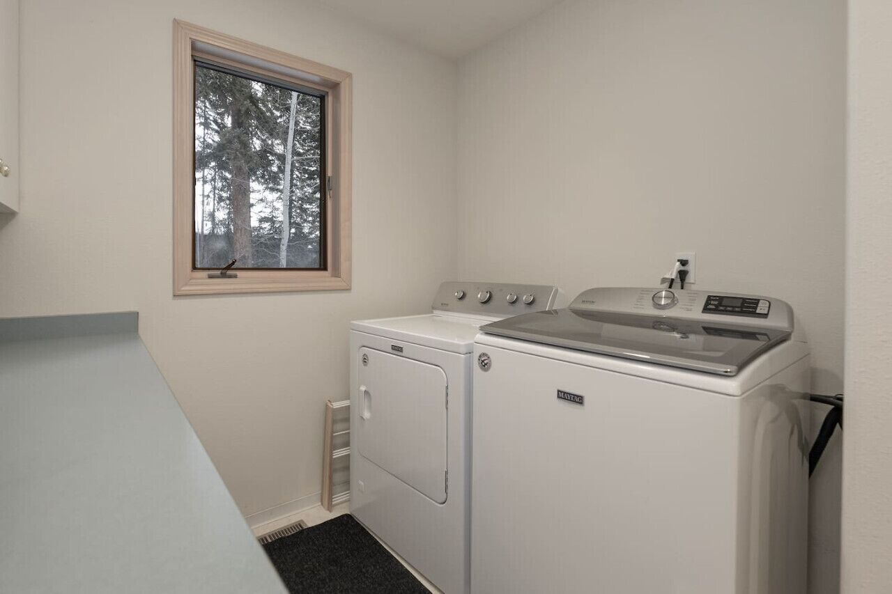 A laundry room with a front-loading washer and dryer, a window with a view of trees, and a small window above the dryer.