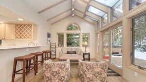 Un interior acogedor con techo alto, barra integrada, zona de estar con otomanas y grandes ventanales con vistas al exterior.