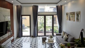 Un salon avec un canapé, une table basse et un téléviseur. Il y a des rideaux, une fenêtre et une plante.
