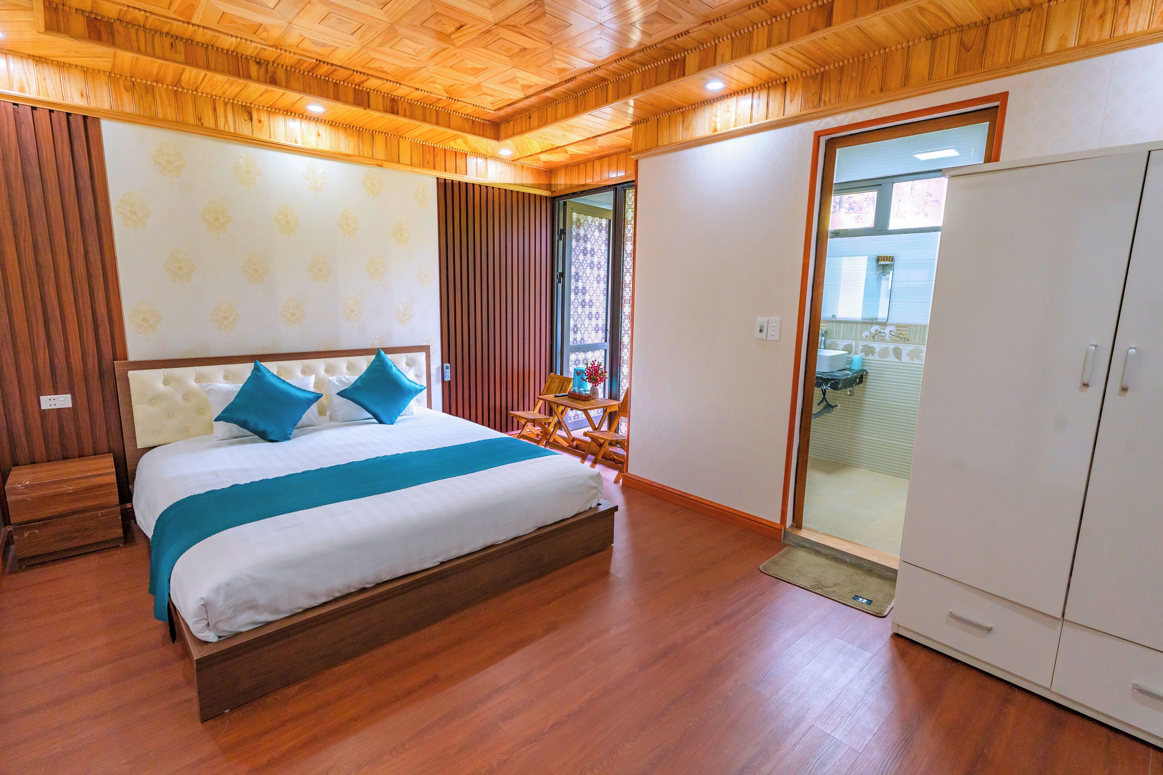 A bedroom with a wooden ceiling, a bed with blue pillows, a wardrobe, and a view through a glass door.