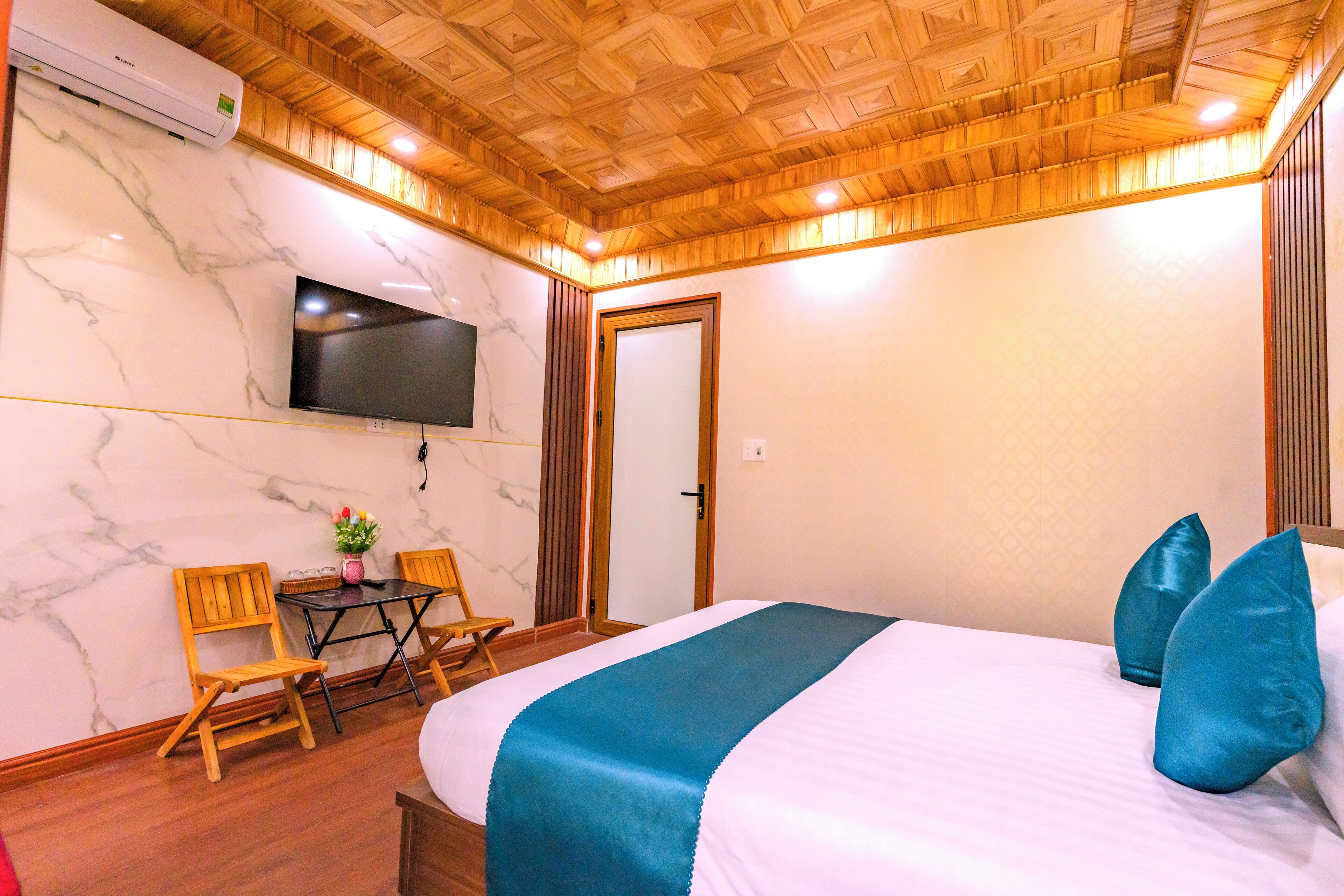 A hotel room with a wooden ceiling, a flat-screen TV mounted on the wall, a bed with blue and white bedding, and a small table with chairs.