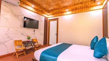 A hotel room with a wooden ceiling, a flat-screen TV mounted on the wall, a bed with blue and white bedding, and a small table with chairs.