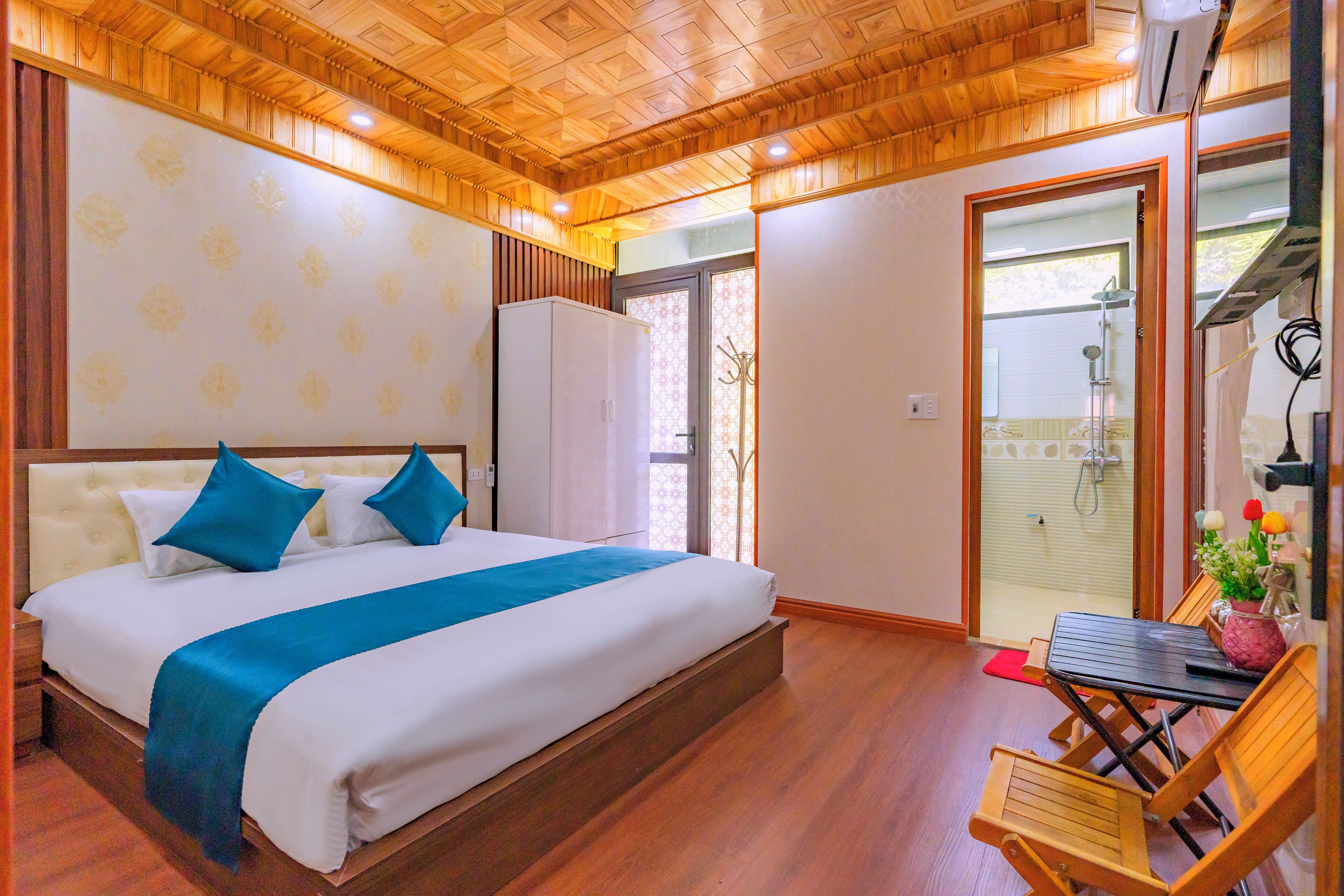 Hotelkamer met een groot bed, houten vloer, een klein tafeltje met stoelen, en een douchecel zichtbaar door een glazen deur.