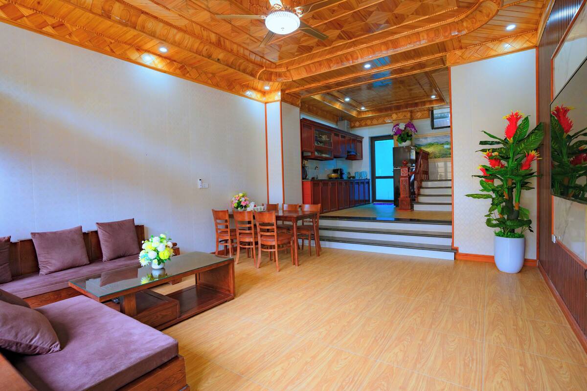 Una sala de estar con sofá, mesa de centro y sillas, techo de madera, y vista a la cocina.