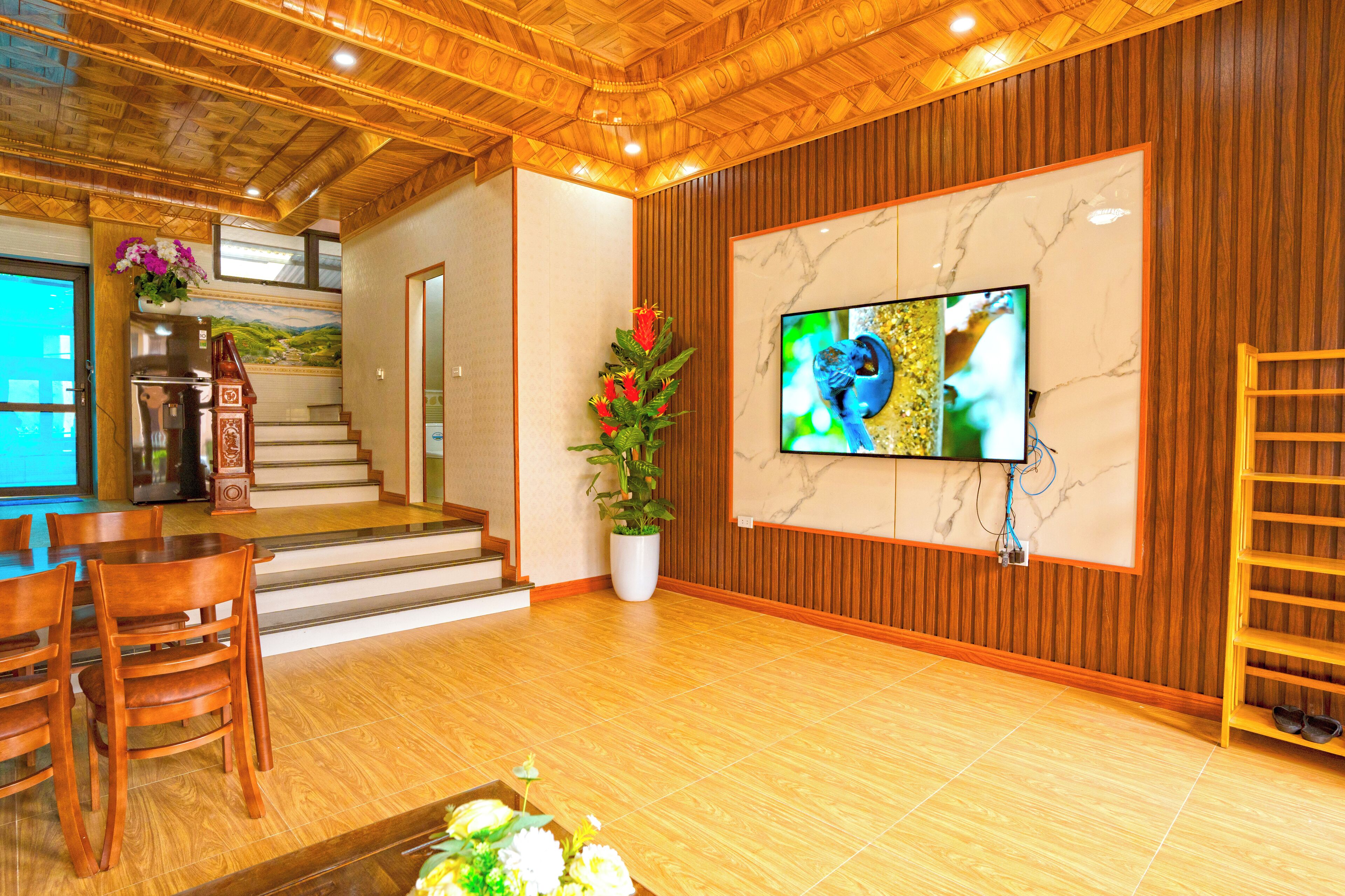 A hotel lobby with wooden flooring, a TV mounted on a marble wall, a staircase, wooden furniture, and a potted plant.