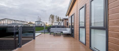En moderne udendørs terrasse med trægulv, en grå sofa og gelænder i glas.