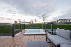 Una terraza de madera con un jacuzzi y un sofá, con vistas a una zona verde y edificios.