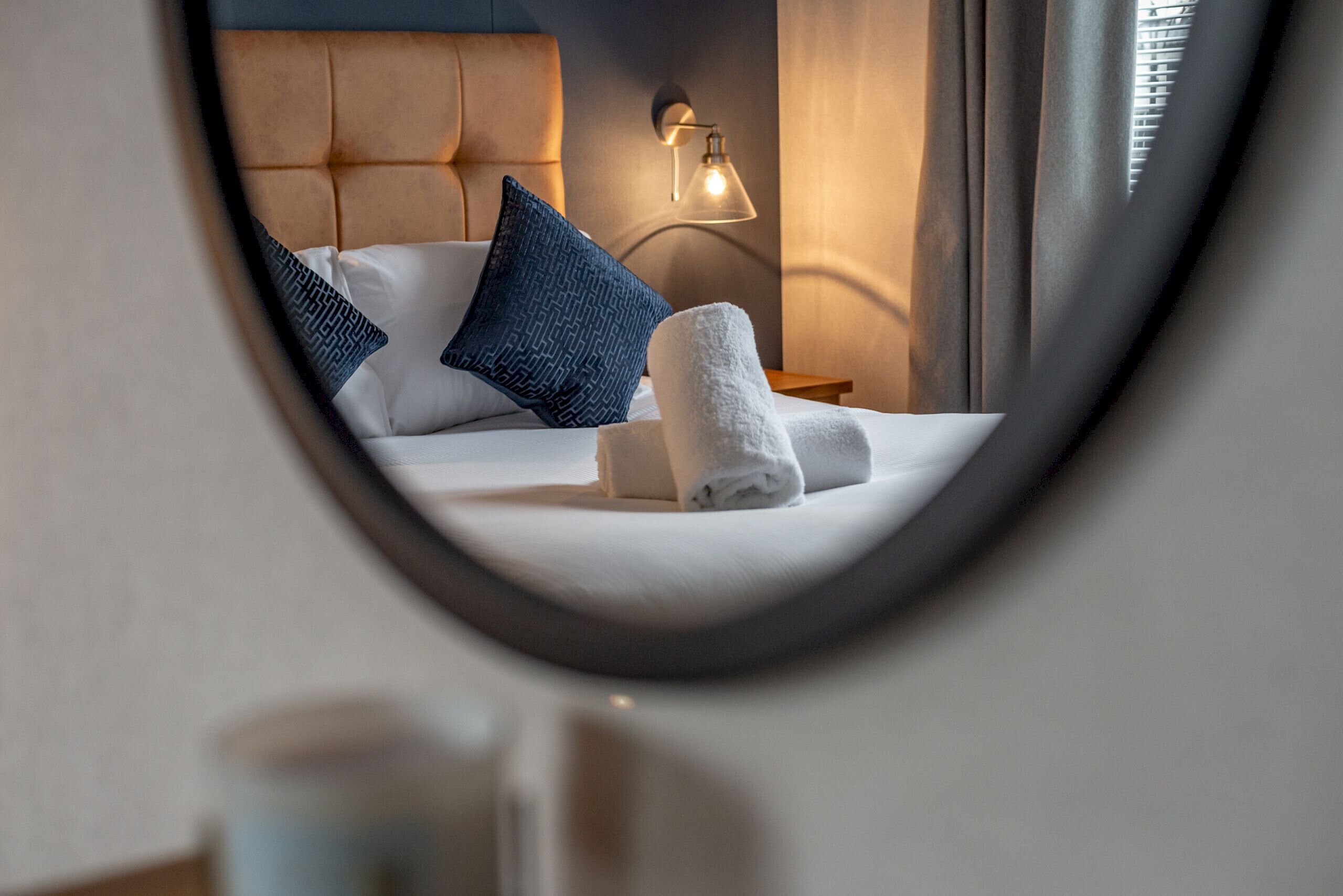 A hotel room with a bed, pillows, a nightstand, a lamp, and a mirror.