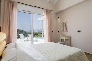 A hotel room with a large bed, a desk, a chair, and a view of a pool and greenery. - Casa di Pietra With 2 Bedrooms and 3 Bathrooms (Kefalonia)