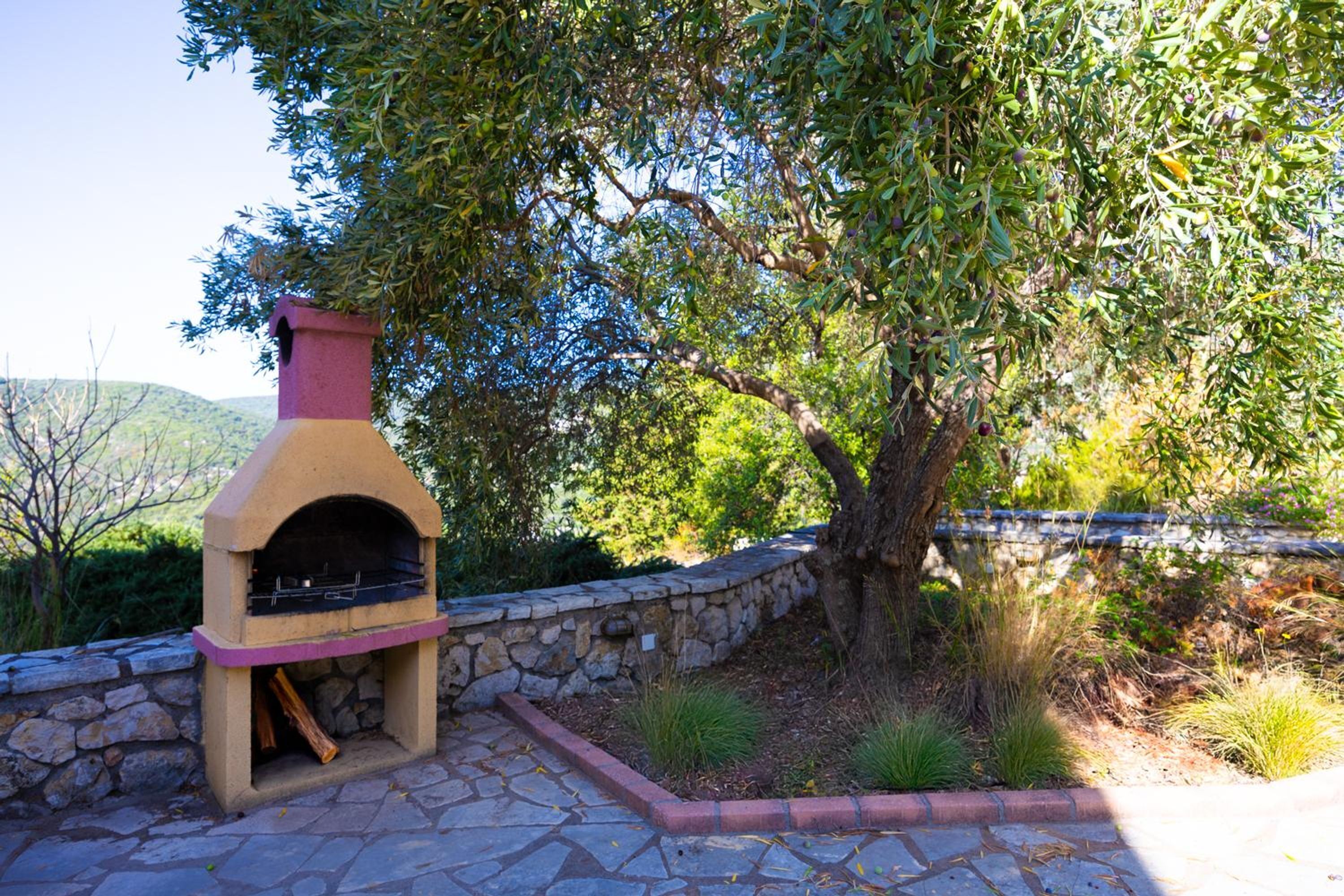 An outdoor stone barbecue grill with a chimney, surrounded by a stone wall and greenery.