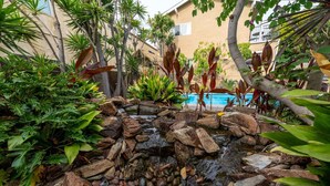A landscaped garden with a small waterfall, surrounded by various tropical plants and a pool in the background. - The OB Hideaway, pool, hot tub (San Diego)