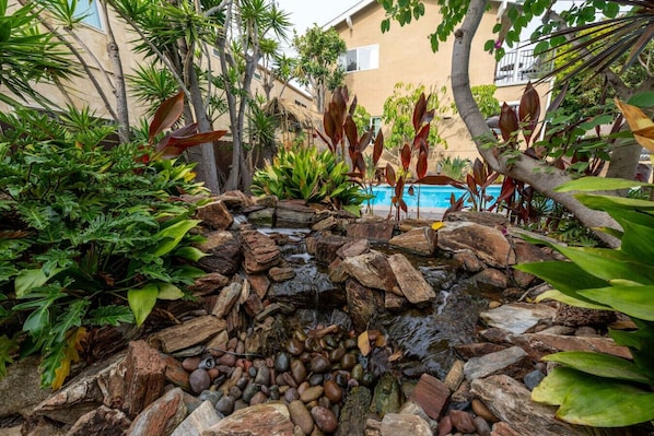 A landscaped garden with a small waterfall, surrounded by various tropical plants and a pool in the background.
