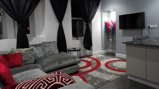 A modern living room with a grey sofa, red and white cushions, a small round table, and a kitchen area with a grey countertop and a flat-screen TV mounted above it. - 2bed Apt Sleeps 4-near Meadows Park, Parking, Wifi (Edinburgh)