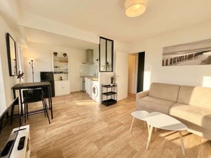 A modern living room with a sofa, a dining table with chairs, a kitchen area with a washing machine, and a large mirror.