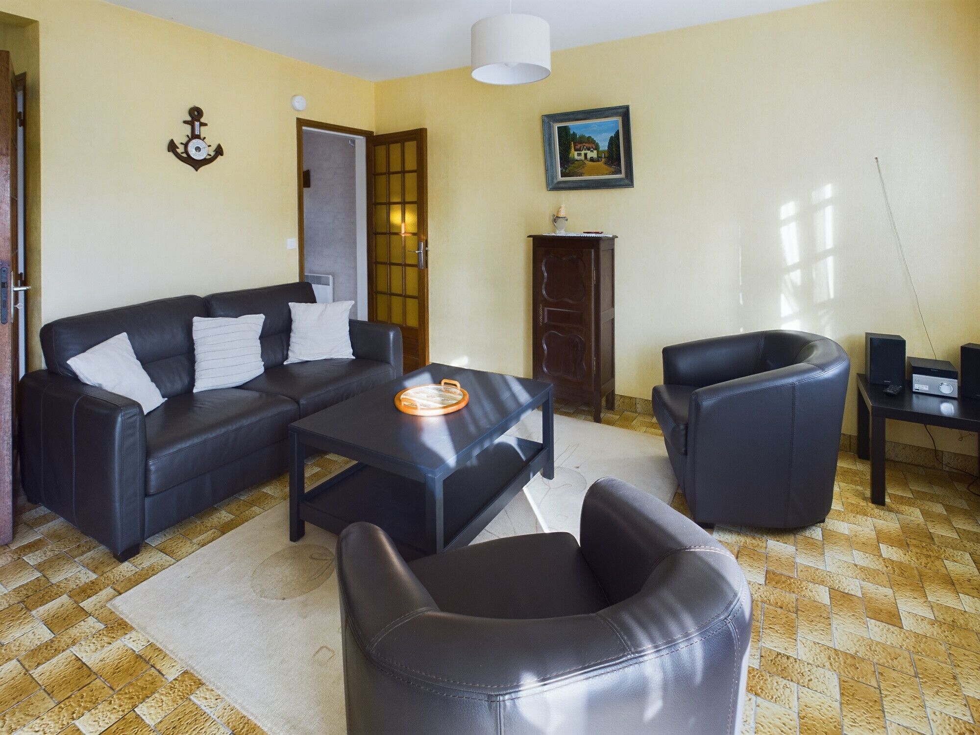 Una sala de estar con un sofá de cuero negro, una mesa de centro y un sillón negro. Hay un cuadro en la pared y una puerta con un panel de vidrio.