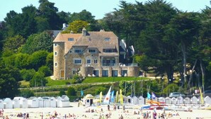 A large stone building with multiple windows and a tower, surrounded by lush greenery and a beach with beach huts. - -HANSEL- Belle Chaumière à Kerascöet (Névez)