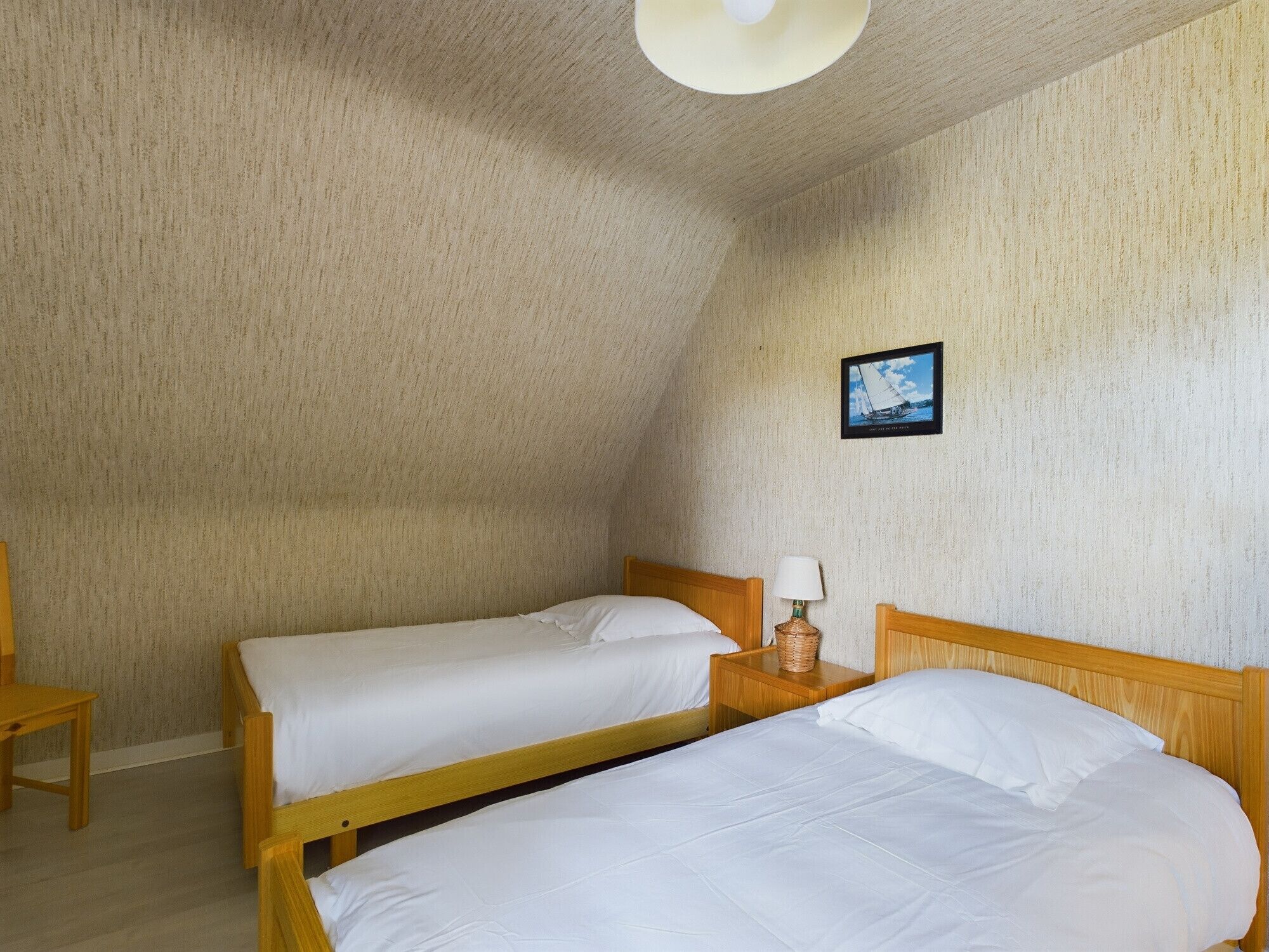 Ein kleiner, gemütlicher Raum mit zwei hölzernen Betten, einem Nachttisch mit Lampe, einem gerahmten Bild an der Wand und einem Holzstuhl.