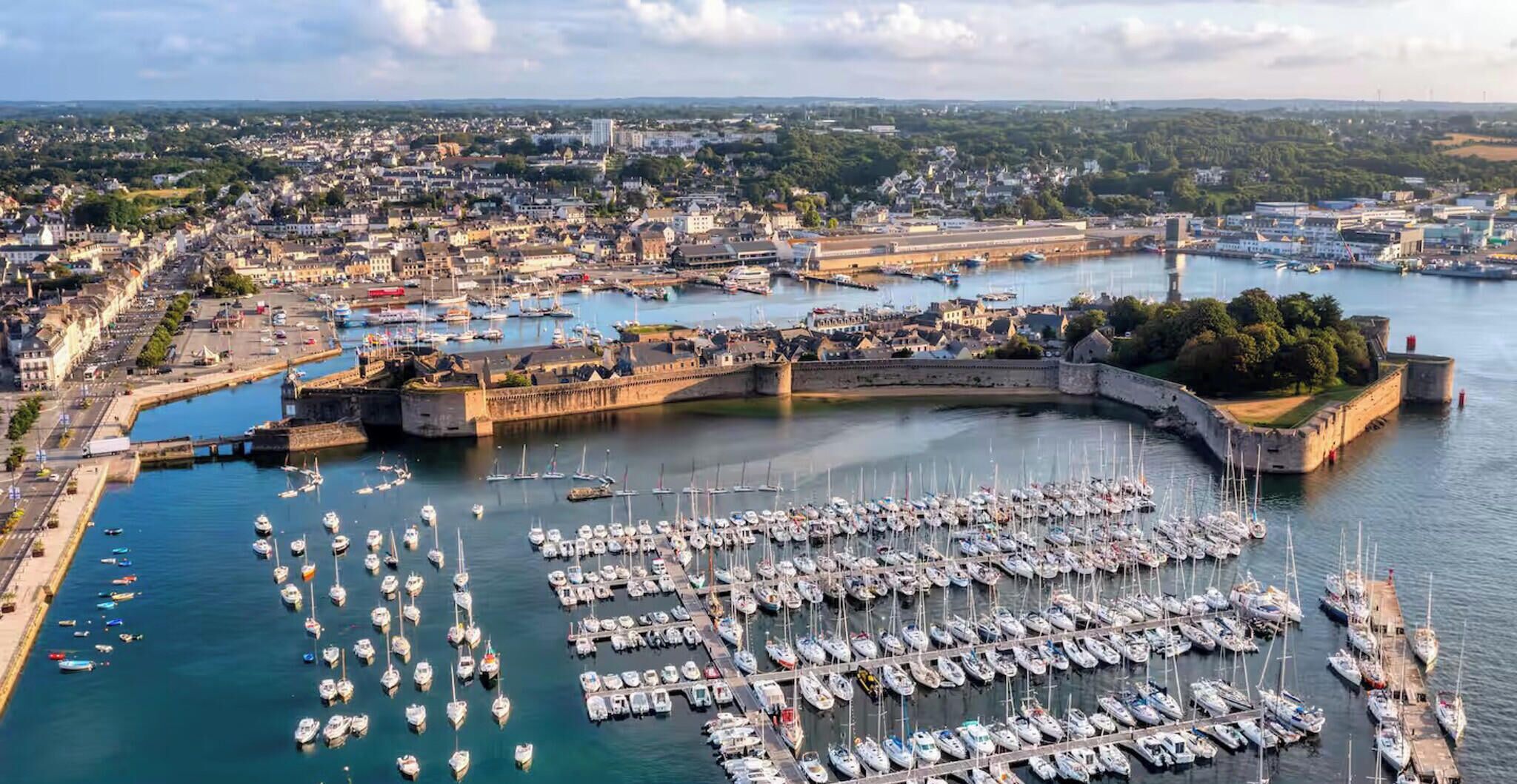 Um porto com vários barcos atracados, uma estrutura fortificada e um horizonte urbano ao fundo.