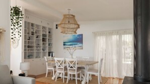 Un comedor con una mesa y sillas blancas, una lámpara colgante y vista al océano a través de cortinas translúcidas.