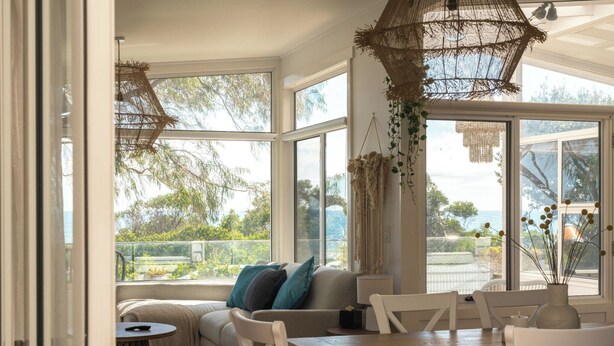 Habitación luminosa con sofá, mesa de comedor y sillas, ventanas amplias con vistas a la vegetación y al mar.