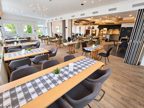 A modern dining area with multiple tables and chairs, a kitchen area, and a view of the outdoors. - Comfortable and versatile room -perfect for relaxation, work and exchange (Winterberg)