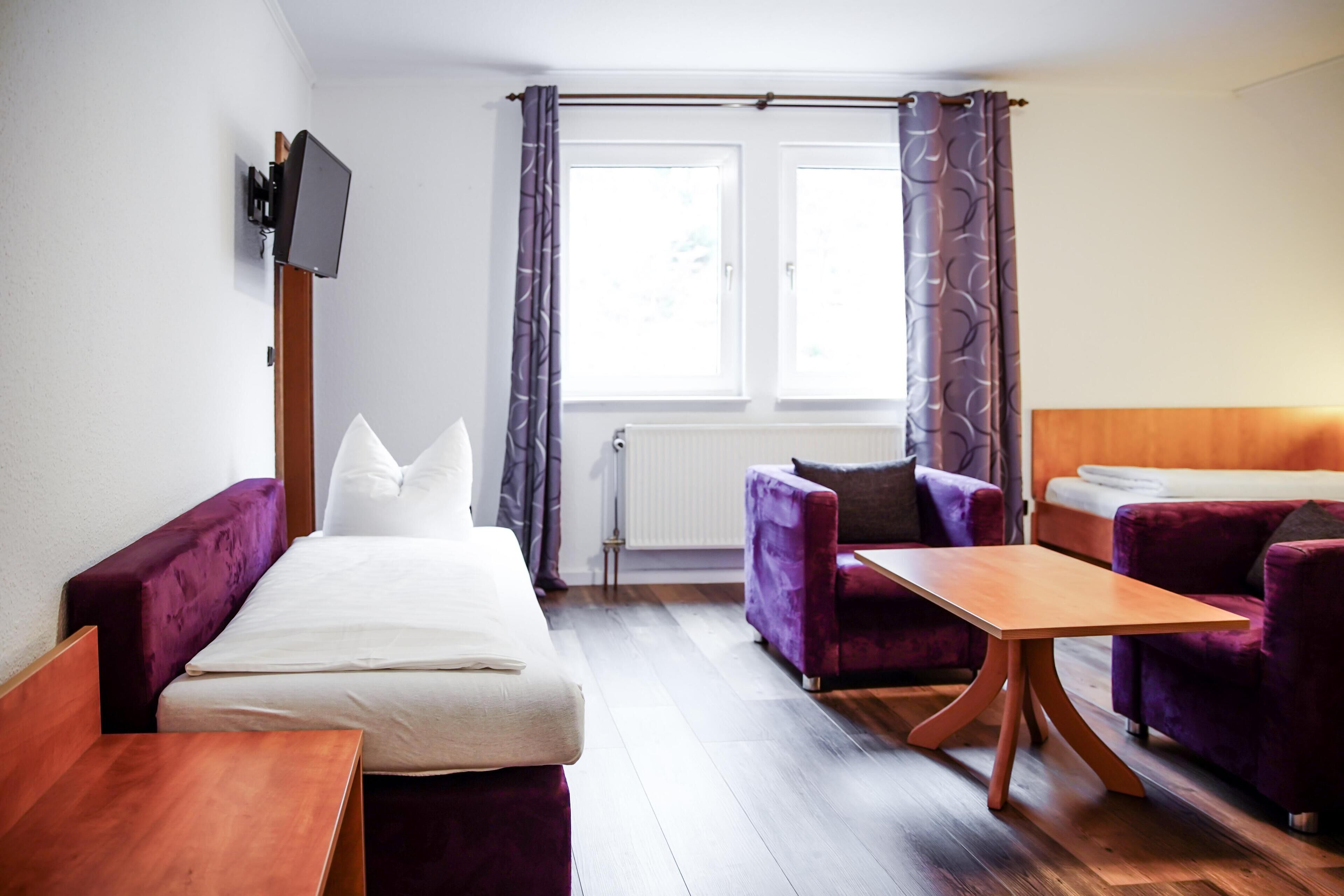 Una habitación de hotel con una cama, dos sillones morados, una mesa de madera, un televisor montado en la pared y una ventana con cortinas.