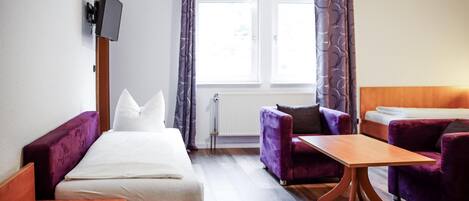 Una habitación de hotel con una cama, dos sillones morados, una mesa de madera, un televisor montado en la pared y una ventana con cortinas.