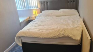 Un lit simple avec une tête de lit rembourrée de couleur grise, une table de chevet avec une lampe et une fenêtre avec des stores.