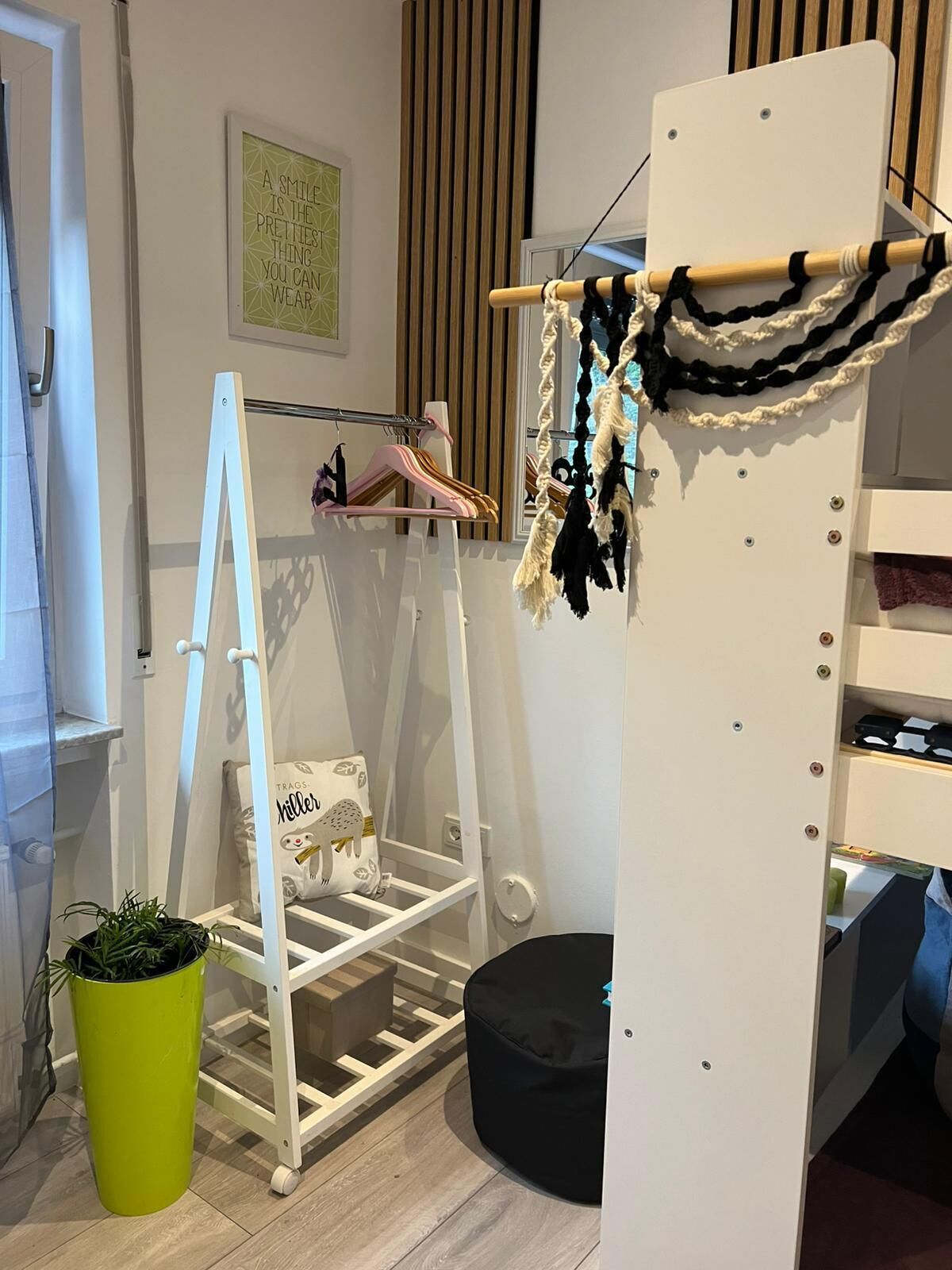 Una habitación con un armario blanco, una percha con ropa, un taburete pequeño y una planta verde.