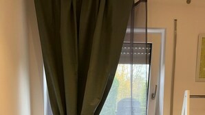 Una habitación con ventana cubierta por una cortina translúcida, un radiador, una planta amarilla y un estante blanco con un cartel.
