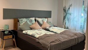 Un dormitorio moderno con una cama grande, una mesita de noche, una lámpara y vistas a palmeras a través de cortinas translúcidas.